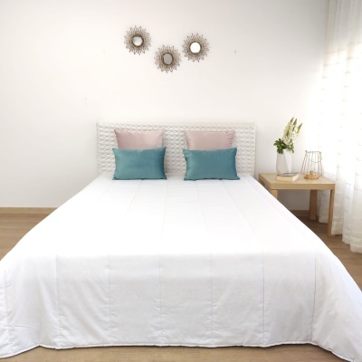 Cama de casal com almofadas azuis e rosas, cabeceira branca acolchoada, mesa de cabeceira com vaso de flores e luminária, três espelhos decorativos na parede
