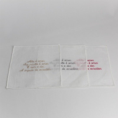 Três panos de tecido branco com bordados em castanho, cinza e rosa com texto em português