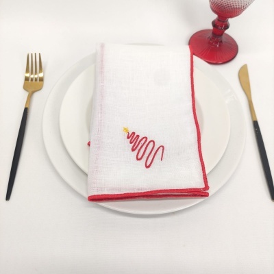 Mesa posta com guardanapo branco bordado, pratos brancos, talheres dourados e pretos, e copo vermelho.