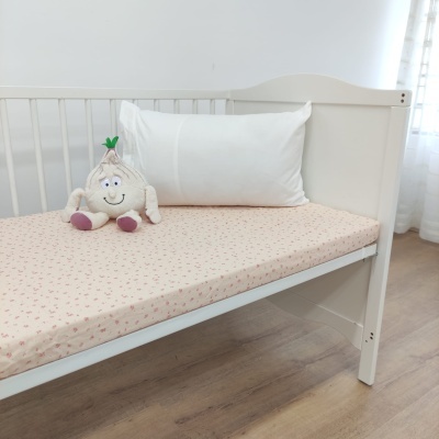 Berço branco com colchão bege com padrão de flores, almofada branca e peluche sorridente em chão de madeira