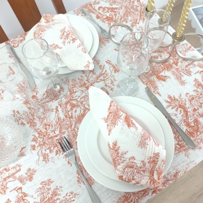 Mesa posta com toalha e guardanapos brancos com padrão em terracota, pratos brancos, talheres metálicos e copos de vidro.