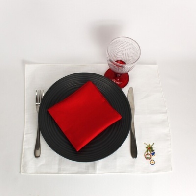 Conjunto de jantar com prato preto, guardanapo vermelho, talheres prateados, copo de vidro com pé vermelho e toalha branca com bordado.