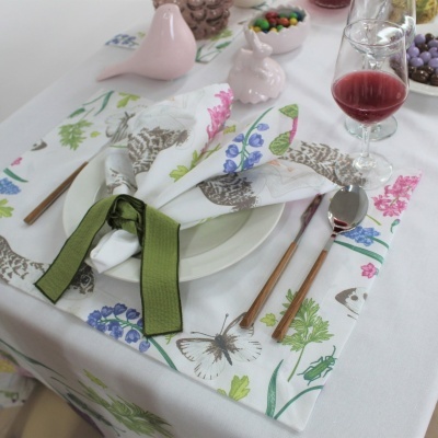 Mesa posta com toalha e guardanapo estampados, talheres com cabos de madeira, copo de bebida vermelha e figuras de pássaros em cerâmica rosa