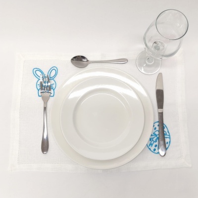 Mesa posta com pratos brancos, talheres de metal e copo de vidro sobre toalha branca bordada com desenhos azuis.