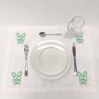 Individual de mesa branco com bordados de coelhos verdes, pratos brancos, talheres e copo de vinho