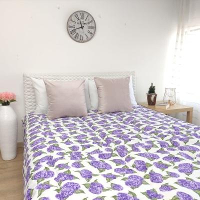 Cama de casal com colcha floral lilás, almofadas brancas e rosa, relógio de parede e mesa de cabeceira simples.