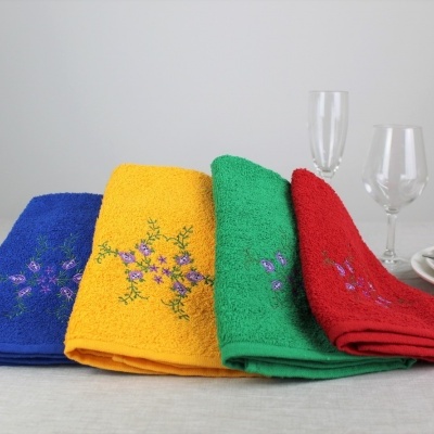 Quatro toalhas coloridas com bordados florais e acessórios de mesa ao fundo