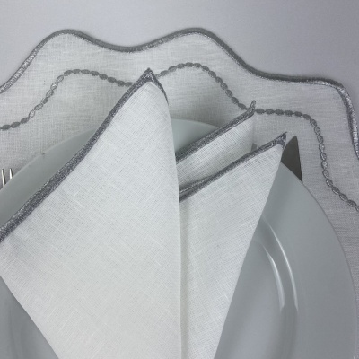 Guardanapo branco com borda cinza sobre prato branco e toalha branca com bordado cinza