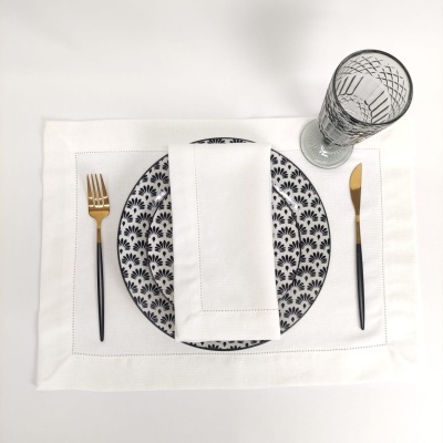 Lugar de mesa com prato branco e preto, talheres dourados e pretos, guardanapo branco e copo de vidro gravado