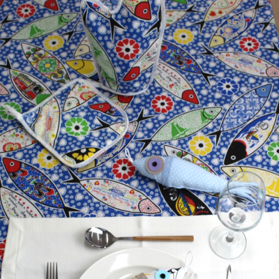 Mesa posta com toalha azul com peixes coloridos e utensílios com padrão de peixe.