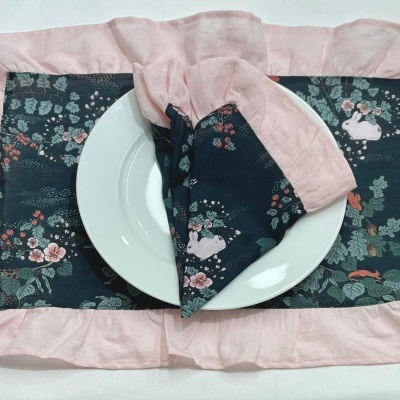 Jogo americano azul escuro com desenhos de flores e coelhos e bordas rosa claro com prato branco e guardanapo combinando