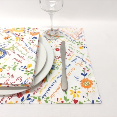 Jogo americano e guardanapo de tecido branco com padrão floral e texto colorido, prato branco, faca e taça de vidro