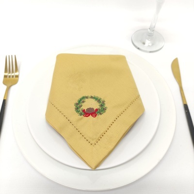 Guardanapo amarelo bordado com coroa de louros e laço vermelho em prato branco, talheres dourados e copo
