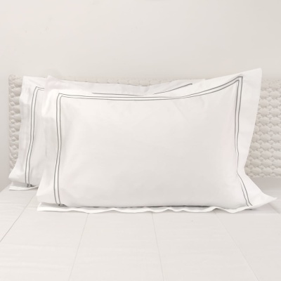 Duas fronhas brancas com duplo risco cinzento na borda sobre cama branca com almofada cinzenta