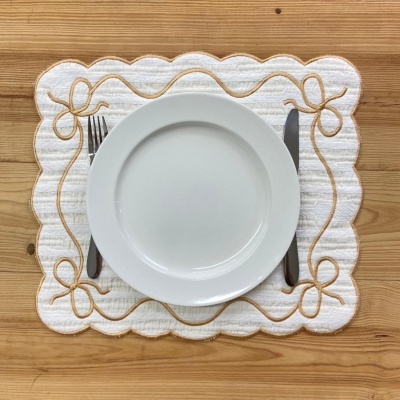 Lugar de mesa branco com bordado dourado, prato branco e talheres sobre mesa de madeira.