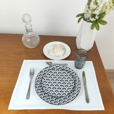 Mesa posta com prato decorado preto e branco, talheres, copo, jarra de vidro, sobremesa e vaso com flores brancas.