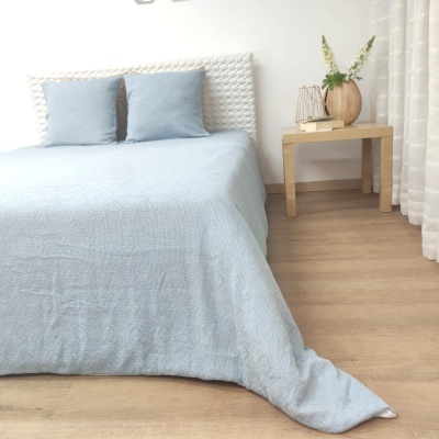 Cama com colcha e almofadas azuis, cabeceira branca, mesa de cabeceira com decoração, chão de madeira.