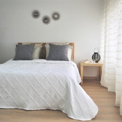 Cama com colcha branca, almofadas cinzentas e madeira clara no quarto com cortinas brancas