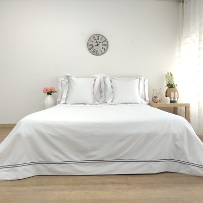Cama de casal com roupa de cama branca e riscas finas azul-escuras, relógio de parede e mesa lateral com flores