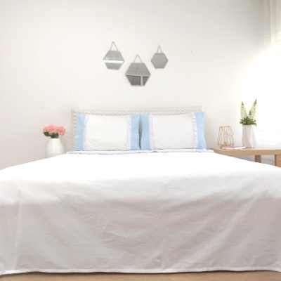 Cama com roupa de cama branca e fronhas azuis e brancas, espeelhos geométricos na parede, vasos com flores ao lado.