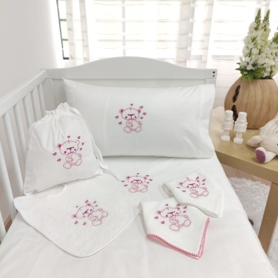 Conjunto de roupas de bebé branco com ursinho rosa bordado numa cama de grades branca
