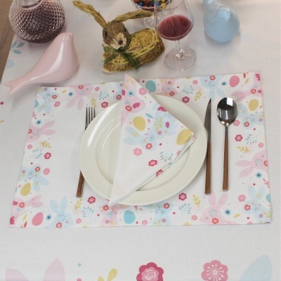 Mesa posta com placemat e guardanapo coloridos de padrão floral e ovos, prato branco, talheres de cabo castanho, copo com vinho tinto e decoração de coelho de páscoa.