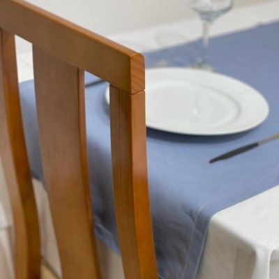 Cadeira de madeira junto a mesa com toalha branca e caminho de mesa azul, prato branco, copo e talher.