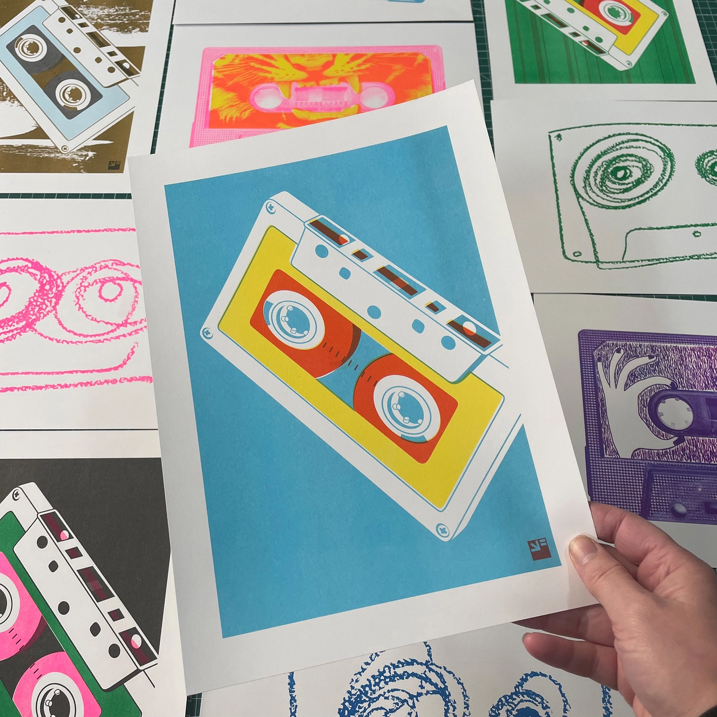 RISO PRINT - CASSETES MISTURADAS - COMBINAÇÃO DE CORES AQUA/AMARELO/VERMELHO - A4 VRIJFORMAAT Impressões artísticas coloridas de fitas cassete em papel branco sobre superfície verde.