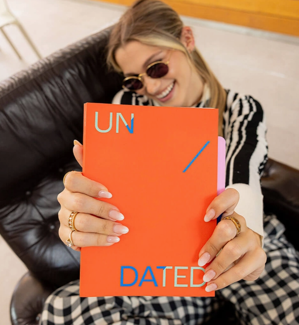 PLANNER A5 "UNDATED" VERMELHO MISHMASH Agenda/caderno laranja com texto UN / DATED nas mãos de uma mulher com unhas de gel e acessórios dourados sentada num sofá preto