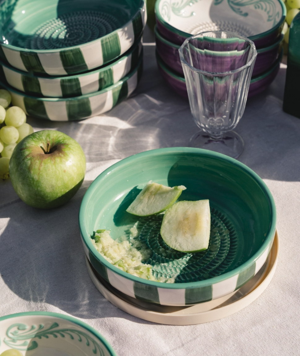 TAÇA RALADOR GRANDE "JADE" ALFAR TIERRA COCIDA Tigelas de cerâmica decoradas com padrões verdes, brancos e roxos e uma taça de vidro transparente numa mesa clara