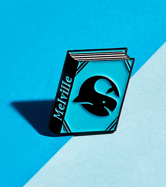 PIN "MOBY DICK BY HERMAN MELVILLE" JUDY KAUFMANN Pin em forma de livro azul e preto com texto 'Melville' e imagem de baleia preta