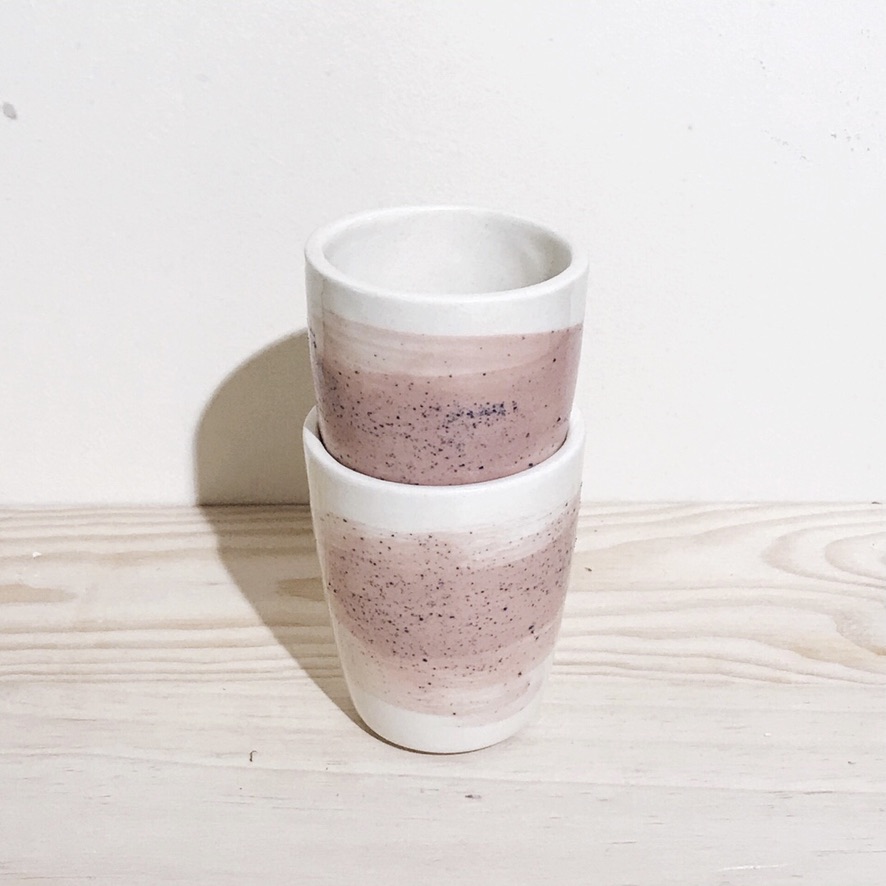 COPO PINCELADO "FREESIA ROSA" MANU SOUZA Dois copos de cerâmica branco e rosa empilhados sobre superfície de madeira