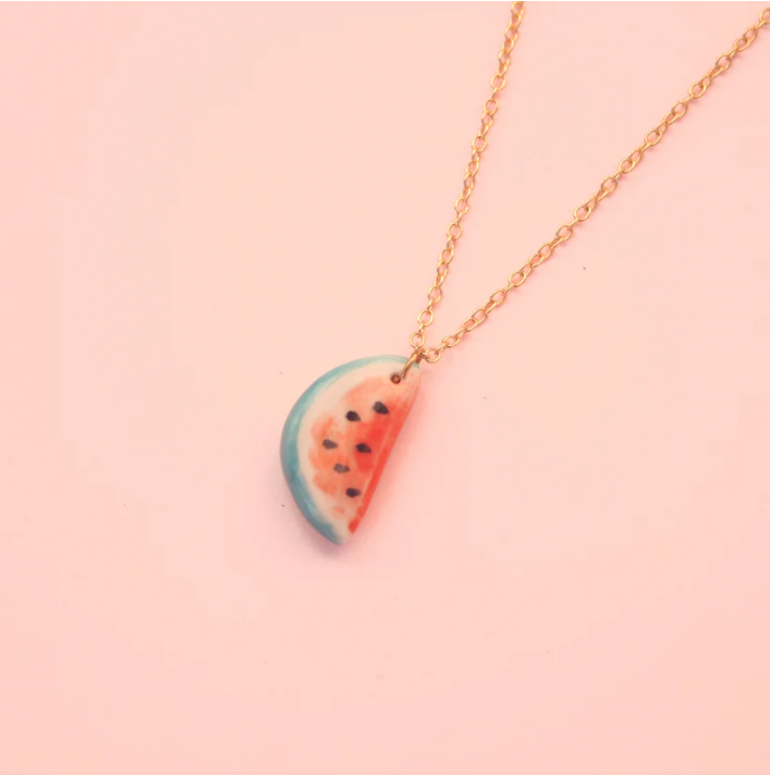 COLAR "WATERMELON" SIAMESE TWIN STUDIO Colar com pendente em forma de fatia de melancia e corrente dourada, sobre fundo rosa claro.