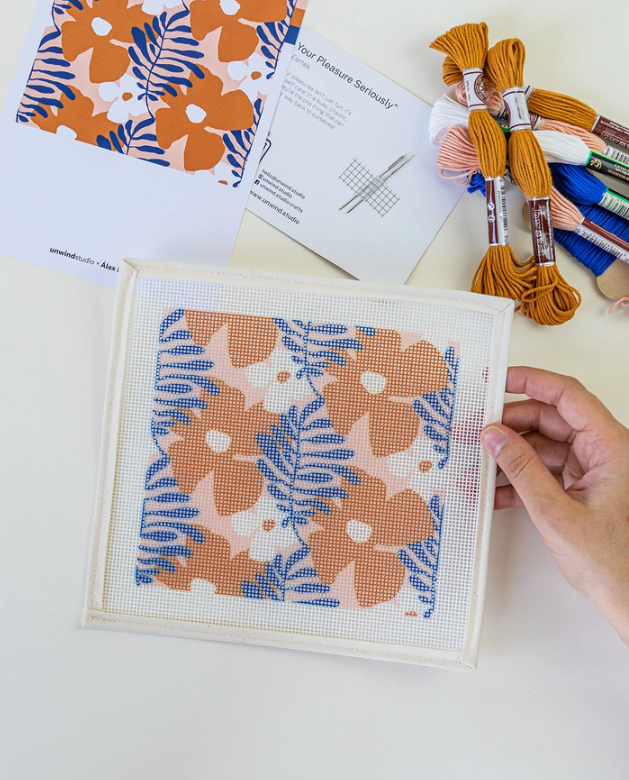 "KIT ESTOJO PARA ÓCULOS DE SOL" ROYAN UNWIND STUDIO Pano de bordado com padrão floral laranja e azul, novelos de linha e folheto ao fundo