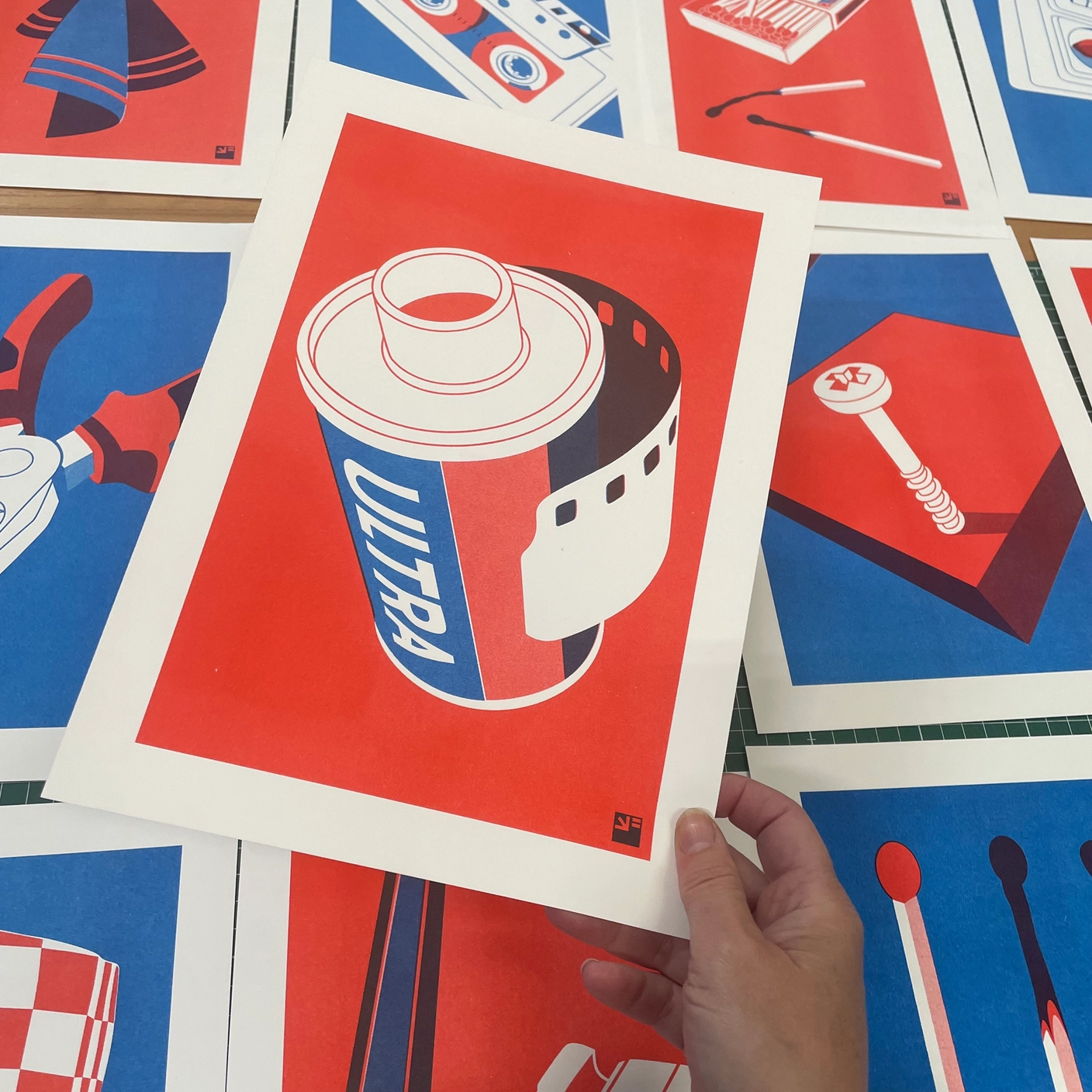 RISO PRINT - ROLO DE FILME PARA CÂMERA ANALÓGICA - EXTRA ORDINARY - A4 VRIJFORMAAT Impressão gráfica colorida de um rolo de filme fotográfico ULTRA segura sobre mesa com outras impressões coloridas
