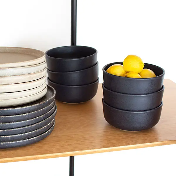 LAPA TAÇA Ø14 BLACK MIDNIGHT C.ª ATLÂNTICA Pratos e tigelas de cerâmica cinza claro e escuro empilhados em prateleira de madeira com limões amarelos nas tigelas.