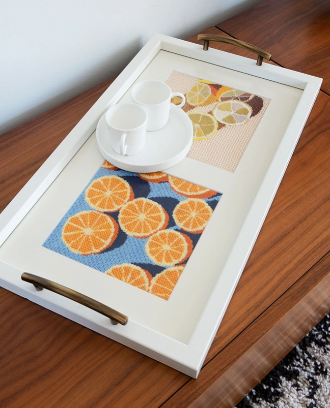 KIT DE PONTO CRUZ "AGRUMES" UNWIND STUDIO Tabuleiro branco com punhos metálicos e painéis coloridos de frutas, sobre mesa de madeira com duas chávenas brancas