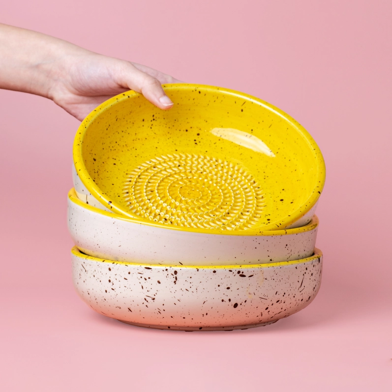 TAÇA RALADOR "CANELA - BRANCO E AMARELO - GRANDE" ALFAR TIERRA COCIDA Três pratos fundos cerâmicos, amarelo e bege com manchas, empilhados sobre fundo rosa, com mão segurando o prato amarelo.