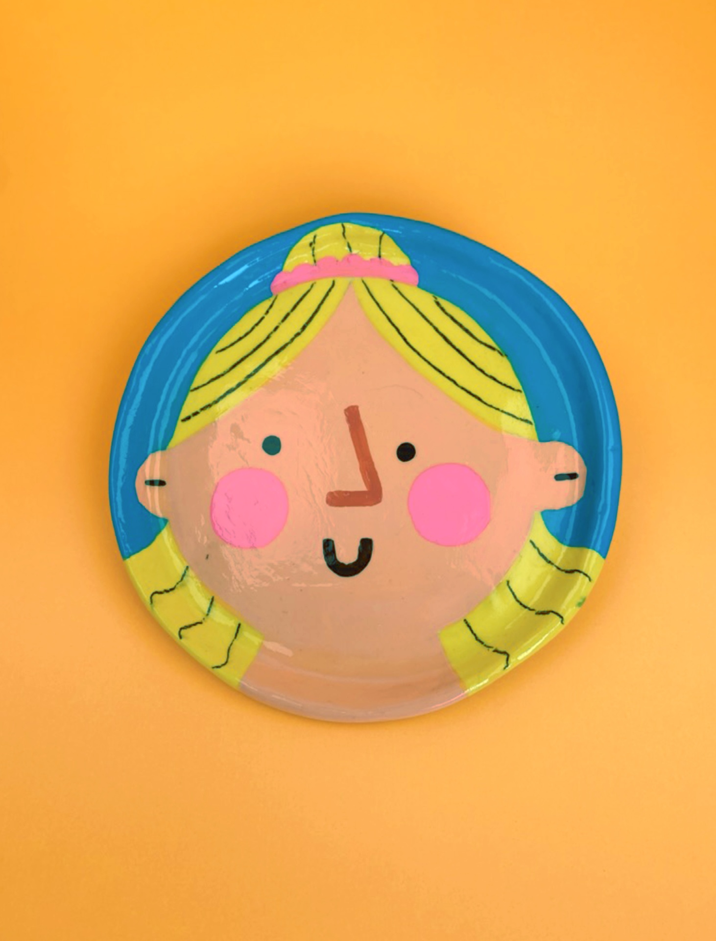 PRATO EM ARGILA "TEODORA" CATPIVARA Prato decorativo redondo com desenho de rosto de criança em fundo azul e amarelo sobre superfície laranja