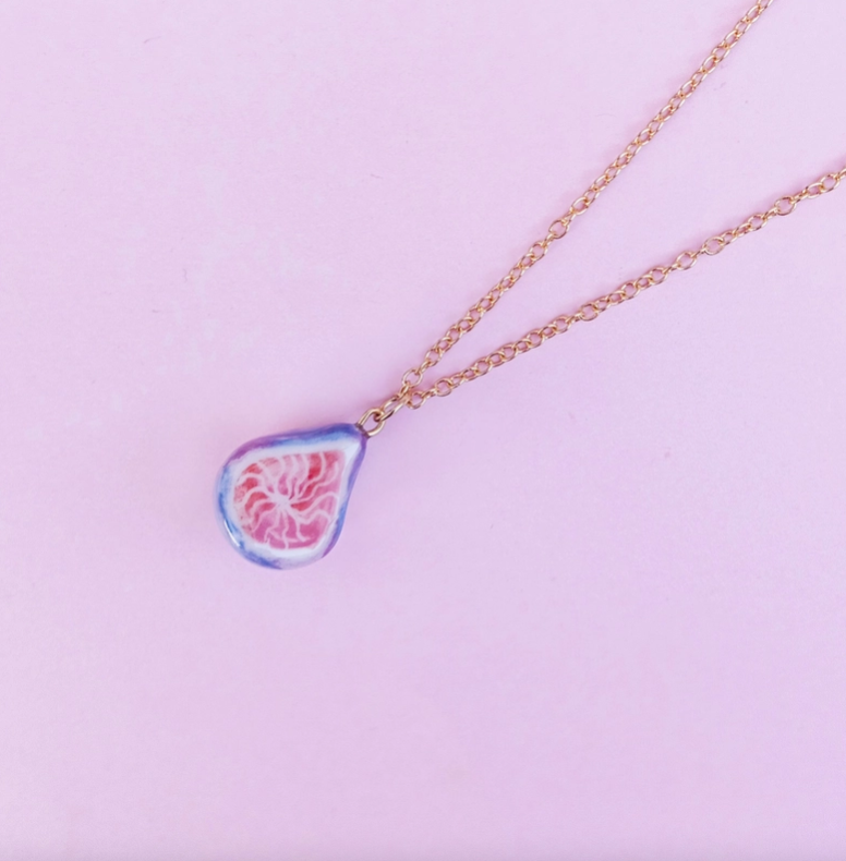 COLAR "FIG" SIAMESE TWIN STUDIO Colar dourado com pendente em forma de gota colorido sobre fundo rosa