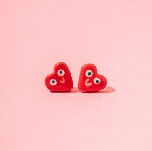BRINCOS CORAÇÃO VERMELHO ANA SEIXAS Brincos vermelhos em forma de coração com olhos e bocas sorridentes sobre fundo rosa