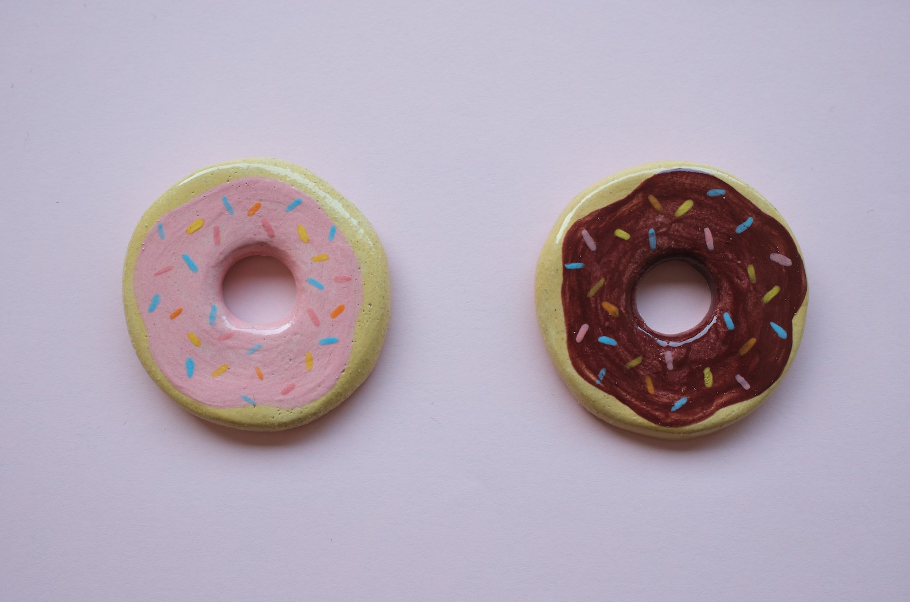 PEÇAS DE CERÂMICA "BREAKFAST FOR DINNER" KIKA Dois donuts de cerâmica pintados com cobertura colorida sobre fundo rosa claro