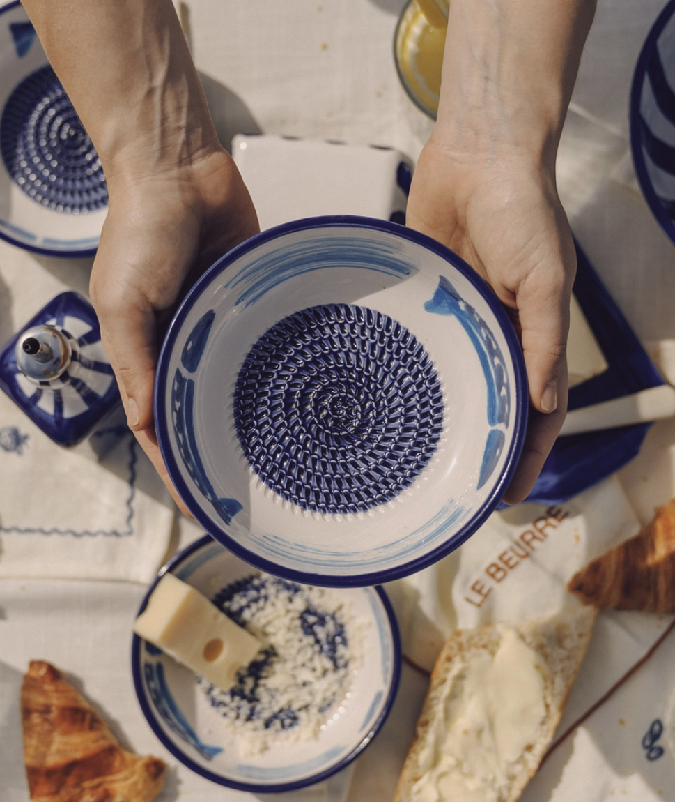 TAÇA RALADOR MÉDIO "ATUM " ALFAR TIERRA COCIDA Tigela cerâmica branca e azul segurada por mãos sobre mesa com queijo, pão e louça azul