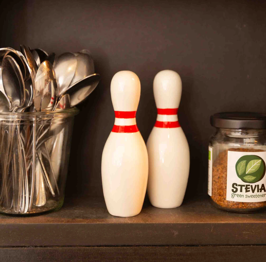 SALEIRO E PIMENTEIRO "BOWLING PINS" DONKEY Saleiros em forma de pinos de bowling brancos com riscas vermelhas numa prateleira com frasco de stévia e recipiente com colheres de metal