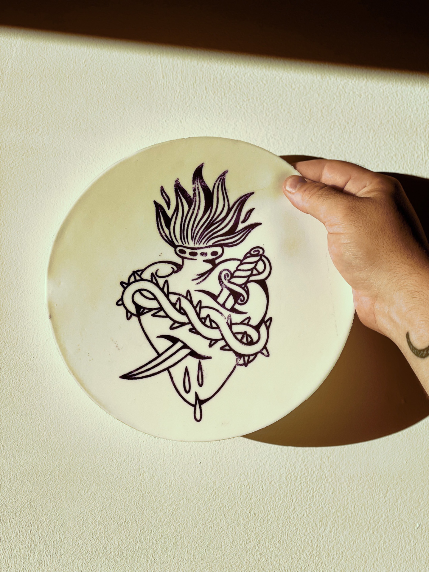STENCIL EM PELE ARTIFICIAL SACRED HEART ZERO TATTOO Prato redondo branco com desenho preto de coração flamejante, faca e vinha espinhosa, segurado por mão