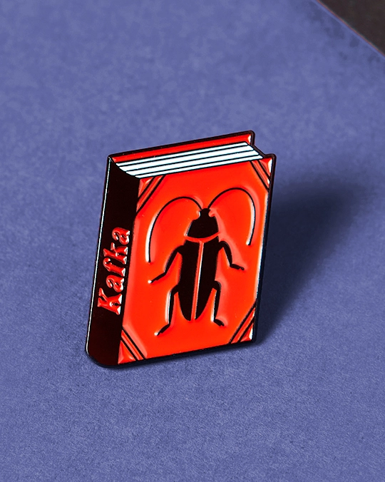 PIN "THE METAMORPHOSIS BY FRANZ KAFKA" JUDY KAUFMANN Pin metálico em forma de livro vermelho com dibujo de inseto e texto Kafka