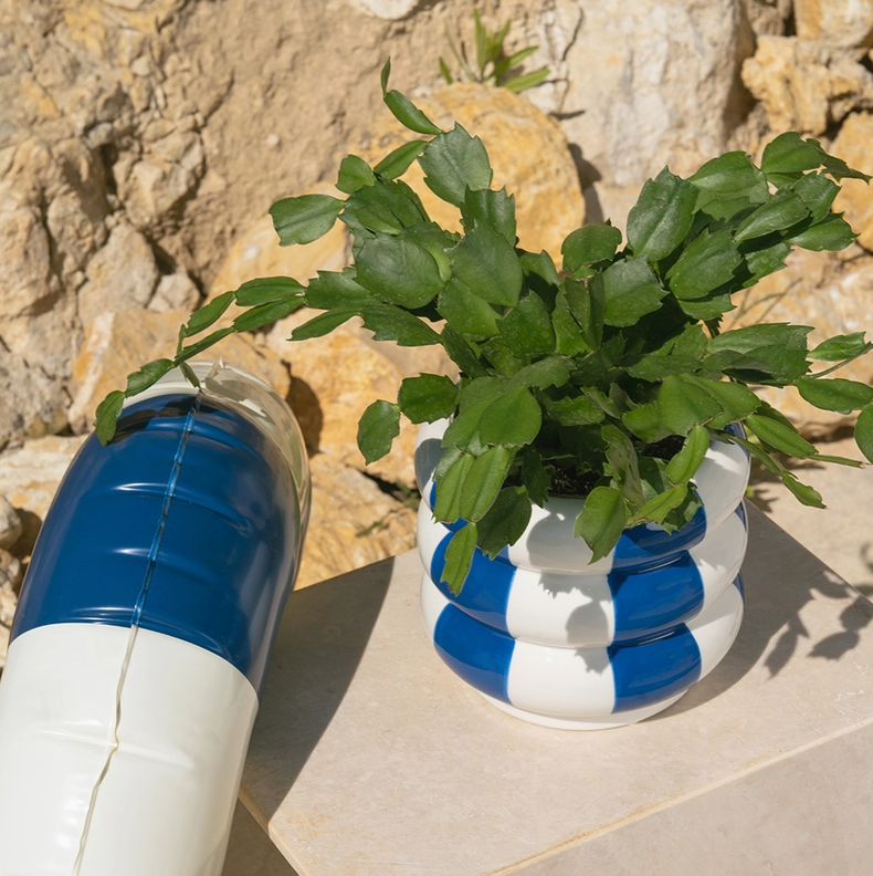 VASO EM CERÂMICA AZUL EM FORMA DE BOIAS EMPILHADAS BALVI Vaso branco e azul com planta verde junto a almofada azul e branca sobre pedra com rochas ao fundo