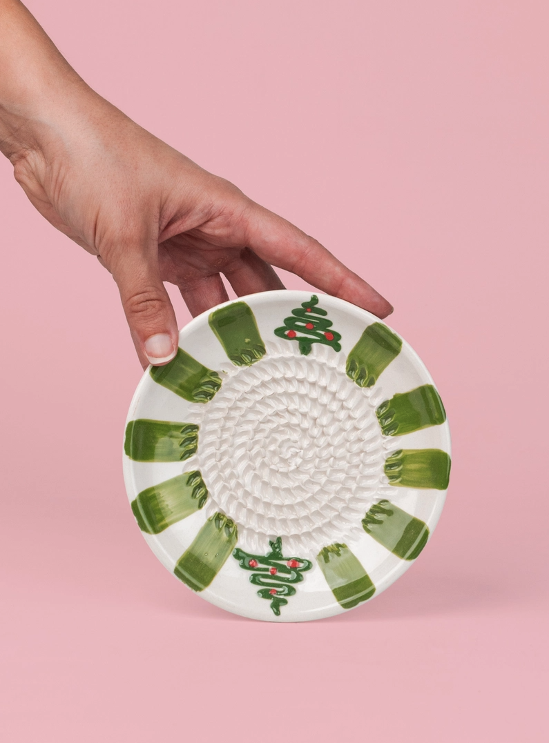 PRATO RALADOR PEQUENO "VERDE PERENE" ALFAR TIERRA COCIDA Prato de cerâmica branco decorado com formas verdes e árvores de Natal, contra fundo rosa.