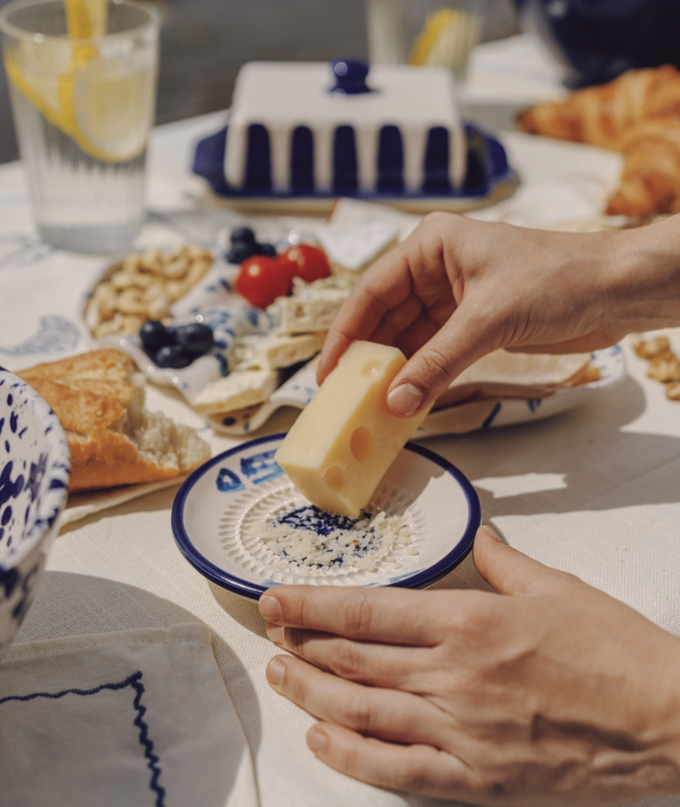 PRATO RALADOR PEQUENO "ATUM" ALFAR TIERRA COCIDA Queijo a ser ralado em prato de cerâmica azul e branca numa mesa com vários alimentos.