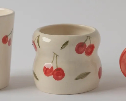 COPO CURVA CITRUS MANU SOUZA Vaso cerâmico creme com padrão de cerejas vermelhas.